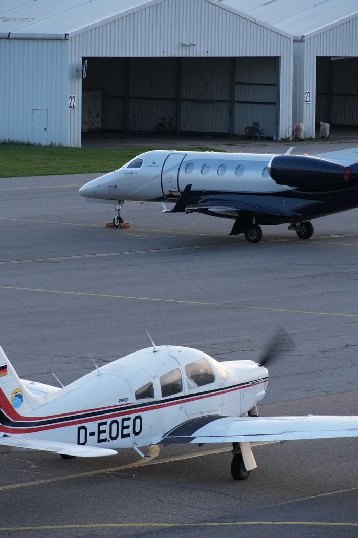Two private aircraft stationed at a European airfield with hangars in the background.