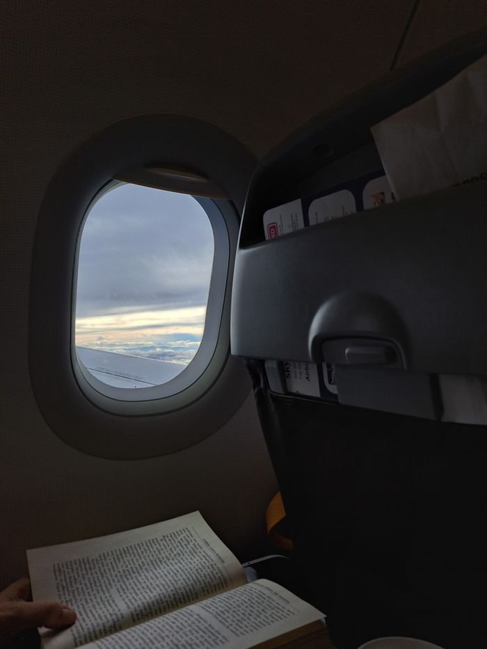 Passenger reading a book on a flight over Thessaloniki with a view out the airplane window.